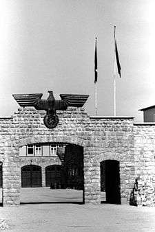 Stone gate topped with a large metal eagle holding a swastika; through the gate a building with two garage doors is visible