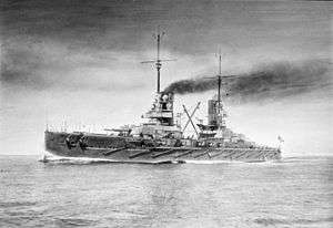 A large warship underway. Dark smoke billows from its smoke stacks.