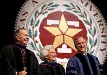 The head and shoulders of three people – an older man, an older woman, and a middle-aged man – wearing formal robes are shown in front of a large circular seal. On the outer edges of the seal the letters "XAS A...IVERSITY...87..." are visible; an inner band of leaves separates the letters from a block T superimposed with a star.