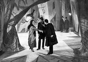 A black-and-white film still.  Three figures stand facing each other on a heavily stylized street set.
