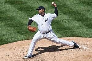 A man in a pinstriped white baseball uniform throws a baseball pitch with his left hand. The breast of his uniform has a navy blue interlocked "NY", with the same logo in white on his navy blue cap. Her is wearing a brown baseball glove on his left hand.