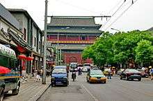 A view up a well-trafficked street to a large dark red building with a Chinese-style roof. On either side are rows of shops. In the foreground is a large metal gantry holding up wires for a trolleybus in the background