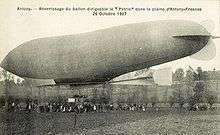 Landscape view with the horizon dividing the photograph in two halves; an airship, looking rather deflated, is settling slightly nose-down in mid-picture foreground at tree-level
