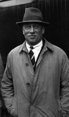 A casual outdoor photo of C. H. Douglas. He is a suntanned man with a white moustache and shrewd smile. He wears a fedora and grubby old gabardine coat.