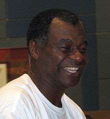 A headshot of a black man. He is looking to the right, wearing a plain white t-shirt and is smiling.