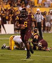 An American football player in a dark red and yellow uniform cradles the football as he sprints towards the camera. Two players in the background lie on the ground.