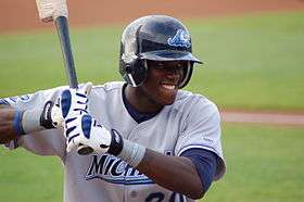 A dark skinned man in a baseball uniform and batting helmet bites his lower lip while holding a baseball bat in a right-handed batting stance.
