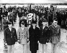 Five men in foreground, four in World War II uniform, one in the center in a suite and overcoat. Behind a color guard stands at attention, with a formation of Filipino American soldiers behind it.