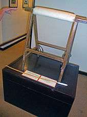A wooden stand of two triangular portions with a padded brace between them on a black pedestal. A hand points from the left, and a small wooden stick with cotton wrapped around one end is in front.