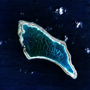 aerial view of a small atoll surrounded by deep blue ocean, a narrow strip of land around a wide area of dappled water in its roughly oblong shaped lagoon.