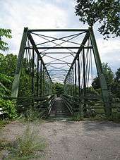 An image of the green Whipple truss bridge from along the side of a road