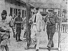 Two Dutch men enter an internment camp, one in a white suit and the other in a military uniform