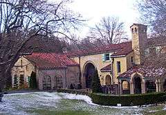 A gold-colored building with a red tiled roof and arches, pavilions and towers. There is an intermittent snow cover on the lawn in front, and two bare trees.