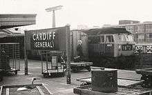 A black-and-white photograph of a railway platform. Two trains stand side-by-side on the image's right while a sign on the platform reads "Cardiff (General)". A formally dressed gentleman stands on the platform, looking away from the viewer. A briefcase, presumably his, rests on a small trolley beside him