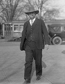 Representative Carl Hayden wearing a dark gray three piece suit and cowboy hat, walking towards the camera and smoking a cigar