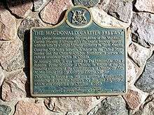 A blue plaque on a stone wall. The plaque has a yellow border, and is mostly rectangular in shape, with the long end oriented horizontally. However, the top side has a camel hump in the centre, with a circle centred at the top of the hump. Inside the circle is an Ontario coat-of-arms. The plaque reads: THE MACDONALD CARTIER FREEWAY This plaque commemorates the completion of the Macdonald-Cartier Freeway (Highway 401), the longest freeway operated without tolls by a single highway authority in North America. Covering 510 miles between Windsor on the Canada–US border and the Ontario-Quebec boundary, it serves the richest economic region in Canada. In January 1965, it was named by The Honourable John P. Robarts, Prime Minister of Ontario, in honour of the two founding architects of the Confederation of Canada, Sir John A. Macdonald and Sir Georges Etienne Cartier. This site is located on the last section of construction, consisting of 15 miles between Ivy Lea and Highway 2, which was completed on October 11, 1968.