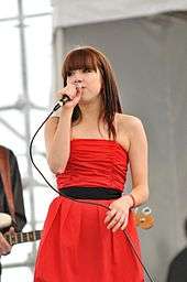 A young woman in long red dress performing live, while handling the microphone in her hand.