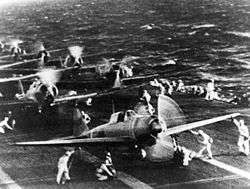 Planes on the deck of an aircraft carrier, with technical crews in white overalls attending the planes.