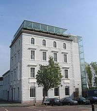 A four-storey building with four bays painted pale grey, with an extra glass storey on the roof and a glass lift on the right side; in front is a small tree and a row of parked cars