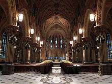 A church interior with a tall vaulted ceilings, wooden pews on a stone floor, and stained glass windows