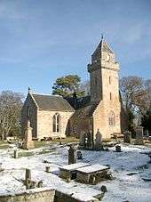 A stone church with a tower in a church yard with grave markers, which is partially covered with snow.