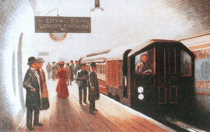 A train pulled by a driving motor car arrives at a station with the driver in view at the controls. Passengers waiting on the platform in Edwardian dress cast long shadows in the brightly lit station.