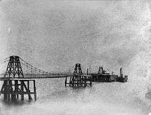 A slightly indistinct black-and-white image of a suspension pier. There is a paddle-steamer docked to the pier at the right. Another one is barely visible through the pier on the left. There is a small building on the platform at the end of the pier, and what may be a crane.