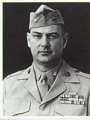 Head and shoulders of a middle-aged white man with a round face wearing a garrison cap and a jacket with four rows of ribbon bars on the left breast and pins on both the lapels and on the collar of the undershirt.