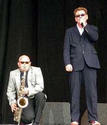 Smash standing onstage, singing into a microphone with Thompson crouched next to him playing a saxophone