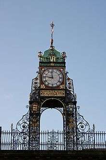 A highly ornate structure in wrought iron, with a railing at the base.  It carries a clock with Roman numerals; this has a red frame bearing the date "1897" in gold, and under it is a plate with an inscription in gold lettering.  Above the clock are the initials "VR" in gold, and at the summit is a green cupola with a weather vane.