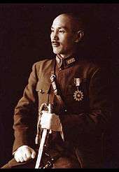A Chinese man in military uniform, smiling and looking towards the left. He holds a sword in his left hand and has a medal in shape of a sun on his chest.