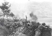 Five Asian soldiers firing weapons and throwing grenades from grass-covered a hilltop. In the middle ground smoke is rising.
