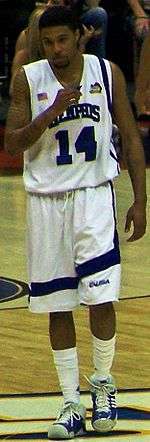 Chris Douglas-Roberts is walking during a college basketball game while wearing the white Memphis University uniform. He is holding a mouthguard and his white socks are almost up to his knees.