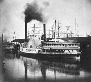 Black-and-white photo of a pair of steamboats moored at a dock; smoke is issuing from one of the boat's funnels; the masts of several sailing ships are visible in the background