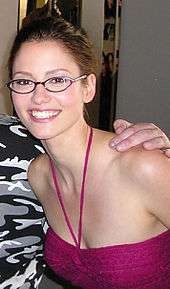 A photo of Chyler Leigh, wearing glasses and a pink T-shirt, smiling at the camera.