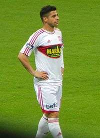 Cicinho on a football pitch, standing with his hands on his hips. He is wearing an all-white kit with red trim