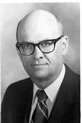 Black-and-white photo of a bald man wearing glasses and a suit with a striped tie