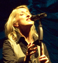 A 44-year old, blonde-haired woman, singing into a microphone with her eyes closed, facing the right of the camera.