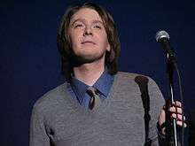 A young white man in a gray pullover with a blue shirt and gray tie, holding a microphone and microphone stand in his left hand, facing upwards.