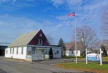 At left, a wooden one-story building with a tall gabled roof. The ground level is white with green trim; the upper level a dark reddish-brown, also with green trim. In the middle is a flagpole with the American flag; behind it is a white church partially obscured by a bare tree. At right is a blue and white sign with "Clermont" prominent on it