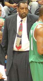 A black person, wearing a black suit and a tie, is standing beside a basketball player in front of the spectators.