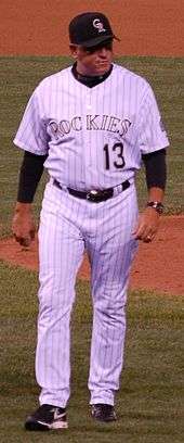 A middle-aged man in a dark baseball cap, white pinstriped baseball uniform with "ROCKIES 13" on the chest, and a black shirt underneath walks away from a pitchers mound.