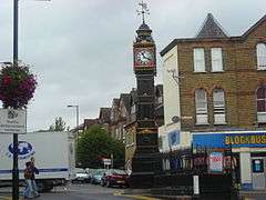 The Clock Tower in South Norwood. It stands on a traffic island. It is a black cast-iron tower, with gilt decoration. The clock face surround is red, with the face being white, with black hands and figures. At the top of the tower is a wind-vane, featuring an arrow and the cardinal compass directions.