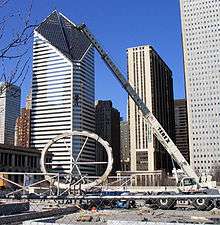 A crane lifts a large, oval-shaped ring in front of several large buildings