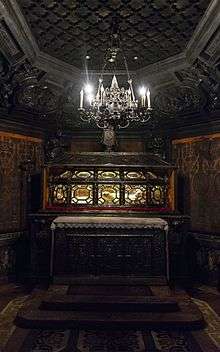 A coffin with glass sides allowing us to see the preserved body within, on a table inside an elaborately carved wooden nook, under a silver electric chandelier