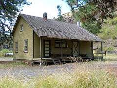 Photograph of a wooden building with a porch/loading dock and steep, rocky slopes in the background.