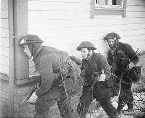 A black and white photograph of three British soldiers advancing beside a wooden house. The lead soldier is carrying a Thompson submachine gun and is peering around the corner of the house, while the other two are carrying rifles.