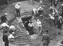Black and white photo of men and women working on constructing an earthen mound with a doorway cut into it. The doorway is lined with sandbags.