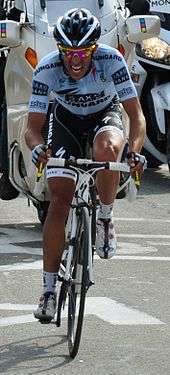 A road racing cyclist in a blue and black jersey with white trim pedaling hard, with a grimace on his face. A motorcycle follows behind.