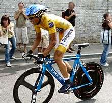 A man with yellow clothes and a blue helmet, riding on a bicycle. In the background some spectators.